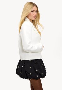 Veste blanche sur mesure avec un col pointu et une coupe structurée, associée à une jupe noire ornée de motifs floraux brodés blancs.