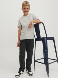 Grå t-shirt med kort ärm och ett fodral, svarta jeans och svartvita sneakers. En blå sliten metallpall står bredvid personen.
