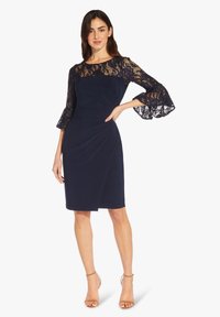 Námořnické šaty s krajkovými rukávy a rozšířenými manžetami, s přiléhavým střihem a diagonálními záhyby v pase. Hladká textura a elegantní design.