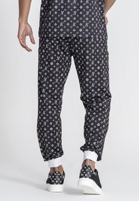 Zwarte joggers met een wit patroon van logo's en symbolen, elastische boorden en een ontspannen pasvorm. Gecombineerd met zwarte sneakers.