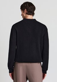 Schwarzer gerippter Strickpullover mit hohem Kragen, langen Ärmeln und lässiger Passform. Das Material wirkt strukturiert und weich.