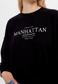 Svart sweatshirt med en vit tryckt grafik som säger "MANHATTAN, UPPER EAST SIDE, NEW YORK," med rund hals och avslappnad passform.