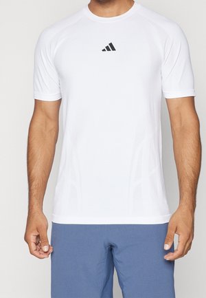 Homme portant un t-shirt de sport blanc à manches courtes avec un petit logo noir sur la poitrine et un short bleu, debout devant un fond uni.