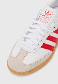 Hvit lær sneakers med røde striper, grå semsket skinndetaljer og hvite lisser. Har en gummisåle i gum og en Adidas-logo på tunga.