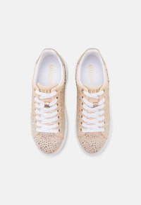 Beige lenkkarit, joissa on teksturoitu pinta, koristeltu strasseilla, valkoiset nauhat ja logo kielekkeessä. Tasainen kumipohja.