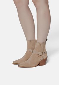 Beige suede ankle boots with a pointed toe, ribbed collar, and adjustable strap with metal accents, featuring a textured heel.