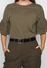 Femme portant un blouse vert olive avec des manches à volants brodées et un pantalon assorti, accessoirisée d'une ceinture noire avec trois anneaux métalliques.