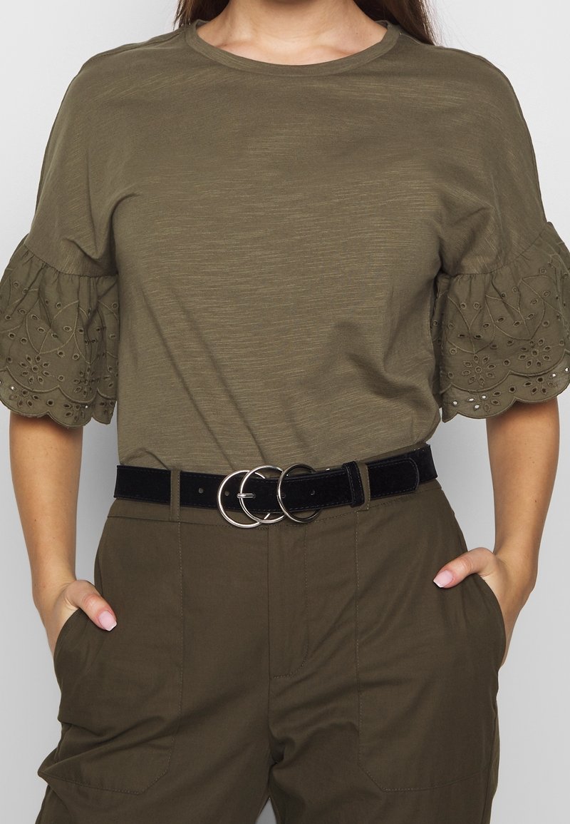 Femme portant un blouse vert olive avec des manches à volants brodées et un pantalon assorti, accessoirisée d'une ceinture noire avec trois anneaux métalliques.