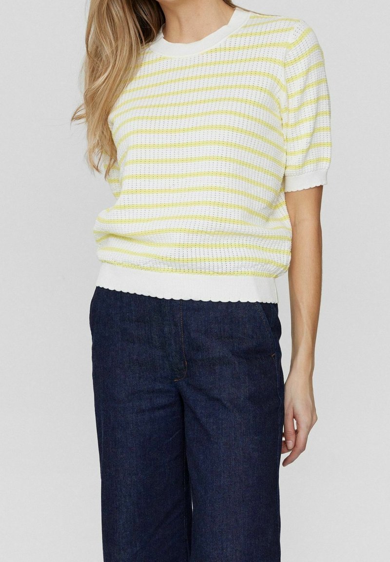 Žena v bílo-žlutém pruhovaném úzkém tričku s krátkým rukávem, kombinovaném s tmavě modrými širokými džíny s vysokým pasem.