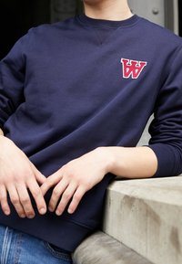 Person som bär en marinblå sweatshirt med en röd "W"-logotyp på bröstet, lutad mot en betongräcke med händerna avslappnat framför sig.