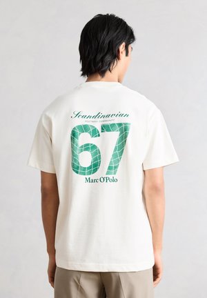 Persona con capelli neri che indossa una maglietta bianca con la scritta verde "Scandinavian Football Community 67 Marc O'Polo" sulla schiena.