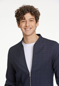 Marinblå zip-up jacka med ett rutmönster, med ståkrage och lätt tyg, bärs över en vit t-shirt.