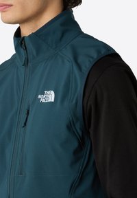 Teal vest med høj krave, lynlås foran og logo detaljer. Båret over en sort langærmet skjorte, der viser en glat tekstur og tætsiddende design.