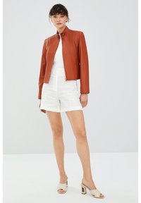 Bruine faux leren jas met een staande kraag, rits aan de voorkant en een gladde afwerking, gecombineerd met witte hooggewaisted shorts en beige hakken.