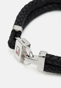 Bracelet en cuir tressé noir avec un fermoir en argent brillant. Le fermoir présente un accent rouge et bleu et le texte gravé "HILFIGER".