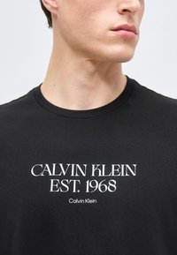 Νεαρό άτομο που φοράει μαύρο μπλουζάκι με λευκό κείμενο «CALVIN KLEIN EST. 1968» στην κεντρική περιοχή του στήθους.