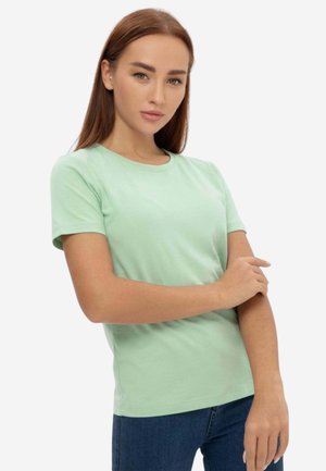 Giovane donna con capelli castani lisci che indossa una maglietta verde chiaro a maniche corte e jeans blu, mentre sistema la manica sinistra.