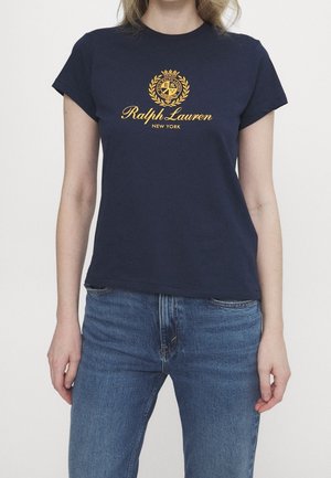 Kobieta w granatowej koszulce marki Ralph Lauren z złotym logo i napisem, w połączeniu z niebieskimi dżinsami, stojąca na tle jednolitego tła.