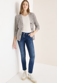 Pletený svetr béžové a hnědé barvy se zapínáním na knoflíky, v kombinaci s přiléhavými modrými džínami a bílými teniskami s světle modrými detaily.