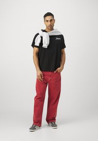 T-shirt noir avec logo en script blanc, pantalon en denim rouge, sweat-shirt gris drapé sur les épaules, baskets grises et blanches.
