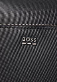 Svart teksturert materiale med en sølvfarget "BOSS"-logo med distinkte bokstavdesign og glidelås langs den øverste kanten.