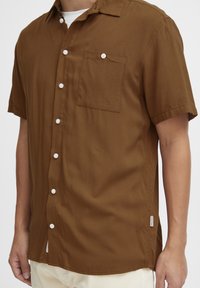 Camisa de botón de manga corta color marrón, hecha de tela suave, con un bolsillo en el pecho izquierdo y botones blancos a lo largo del frente.