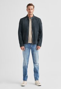 Schwarze Lederjacke mit Stehkragen, Reißverschluss vorne und zwei Seitentaschen, kombiniert mit hellblauen Jeans und cremefarbenen Turnschuhen.