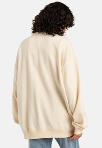 Overmaat crèmekleurige sweatshirt gemaakt van zachte stof, met een verlengd schouderdesign, geribbelde manchetten en een afgeronde zoom. Geen zichtbare logo's of patronen.