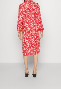 Robe à motif floral rouge avec de longues manches bouffantes, un col montant et une longueur midi. Le tissu semble léger et légèrement transparent.