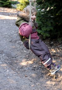 Gyermek egy fából készült hintán, zöld és rózsaszín színű, szőrme díszítésű szigetelt kabátot, barna vízálló nadrágot és barna túracipőt visel kiemelésekkel.