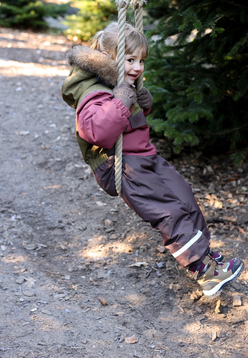 Gyermek egy fából készült hintán, zöld és rózsaszín színű, szőrme díszítésű szigetelt kabátot, barna vízálló nadrágot és barna túracipőt visel kiemelésekkel.