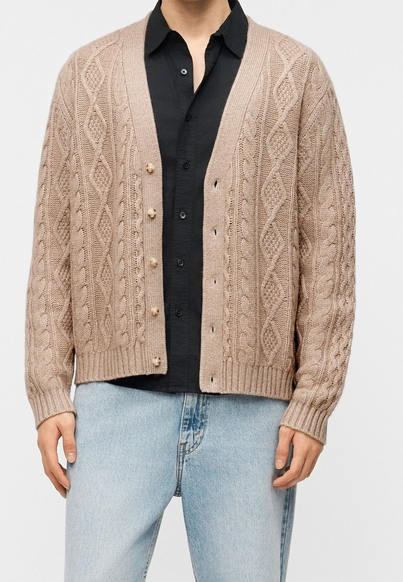 Cardigan beige en maille torsadée avec boutons en perle porté sur une chemise noire à col et un jean bleu clair.