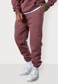 Bordeauxfarbene Jogginghosen aus weichem Stoff mit elastischen Bündchen, seitlichen Taschen und einem Logo-Detail. Kombiniert mit weißen Sneakern.
