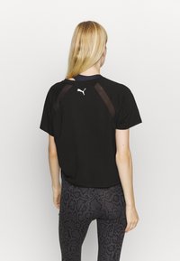 Kvinna med långt blont hår iförd en svart Puma-t-shirt med nätdetaljer och mörka mönstrade leggings, stående vänd bort mot en enfärgad bakgrund.