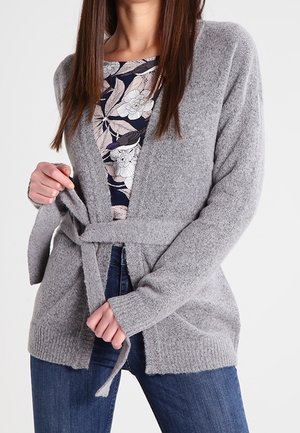 Mujer con cárdigan gris con cinturón sobre un top azul marino con estampado floral y vaqueros azules, ajustándose el cinturón del cárdigan en la cintura.