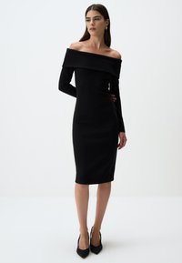 Abito nero senza spalle con maniche lunghe, design aderente, scollatura a coste, lunghezza fino al ginocchio, abbinato a scarpe nere con tacco alto.