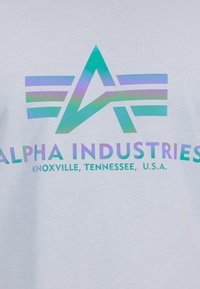 Camiseta blanca con un diseño gráfico iridiscente que presenta una forma abstracta de "A" y elementos a rayas, con el texto "ALPHA INDUSTRIES KNOXVILLE, TENNESSEE, U.S.A."