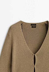 Beżowy sweter robiony na drutach z głębokim dekoltem w kształcie V, prążkowaną teksturą oraz zapinany na guziki z przodu. Otwory po bokach zapewniają wentylację, a luźny krój dodaje swobody.