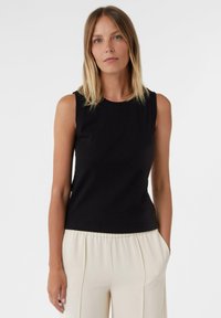 Schwarzes ärmelloses Top aus geripptem Material mit Rundhalsausschnitt, kombiniert mit hellen, plissierten Hosen, vor einem schlichten Hintergrund.