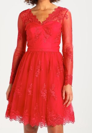 Mujer con un vestido rojo hasta la rodilla, bordado con flores de encaje, mangas largas transparentes y un escote y dobladillo con borde en forma de ondas.