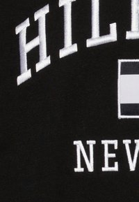Black fabric with white embroidered text reading “HILFIGER” above “NEW YORK.” Includes rectangular logo with red, white, and blue stripes.