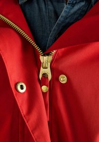 Czerwona kurtka częściowo rozpięta z złotym zamkiem błyskawicznym i zatrzaskami na niebieskiej dżinsowej koszuli.