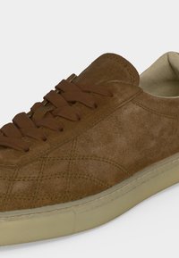 Καφέ σουέντ αθλητικό παπούτσι με ταιριαστά κορδόνια, ορατή ραφή και μπεζ καουτσούκ σόλα πάνω σε ανοιχτό φόντο.