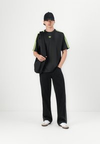 Sort oversized T-shirt med neongrønne striber, sorte baggy bukser, hvide og tan sko samt en sort kasket. Enkel design med logo accenter.