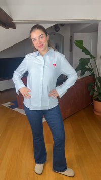 Femme debout à l'intérieur, les mains sur les hanches, portant une chemise rayée bleu clair avec un patch en forme de cœur rouge, un jean bleu et des chaussures beiges.