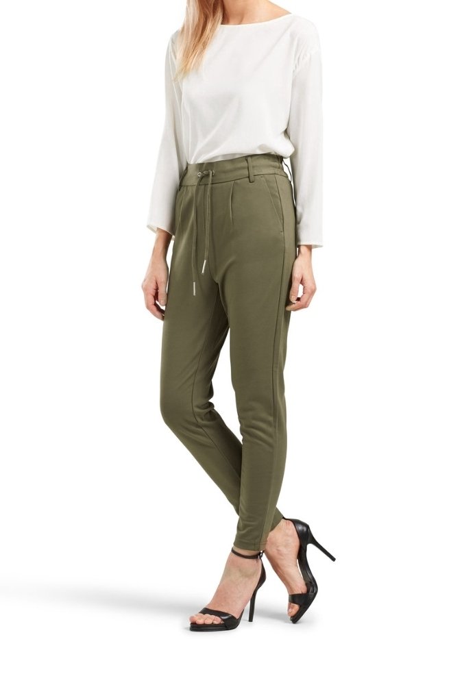 Femme portant un pantalon slim vert olive avec taille à cordon, associé à un haut blanc à manches longues et des sandales noires à talons hauts.