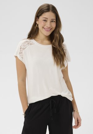 Junge Frau mit langen braunen Haaren lächelt, trägt ein weißes Top mit Spitzenschultern, schwarze Hosen mit Kordelzug, goldene Creolen und Armbänder.