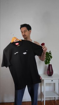 Man die een zwart Diesel T-shirt aan een hanger vasthoudt, staand naast een witte tafel met een paarse vaas waarin een groen blad en een rode roos zitten.