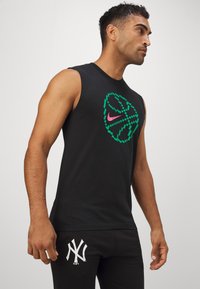 Melnā bezpiešu sporta krekls ar daudzkrāsainu ģeometrisku Nike logo; valkāts ar melniem biksēm, kuru sānos ir balts NY logo.
