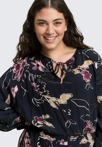 Navy bloemenblouse met lange mouwen, voorzien van een striknek en een elastische tailleband. Het patroon bevat roze en beige bloemen met groene accenten.